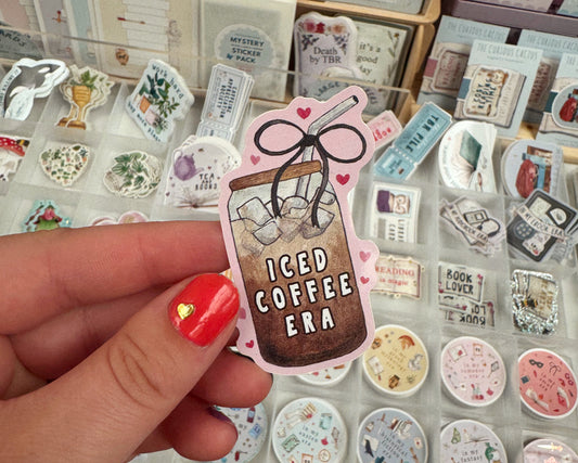 A hand with red nail polish holds a die-cut sticker featuring an iced coffee jar with the text 'ICED COFFEE ERA' and small pink hearts. The background shows a display of various decorative stickers and small items.