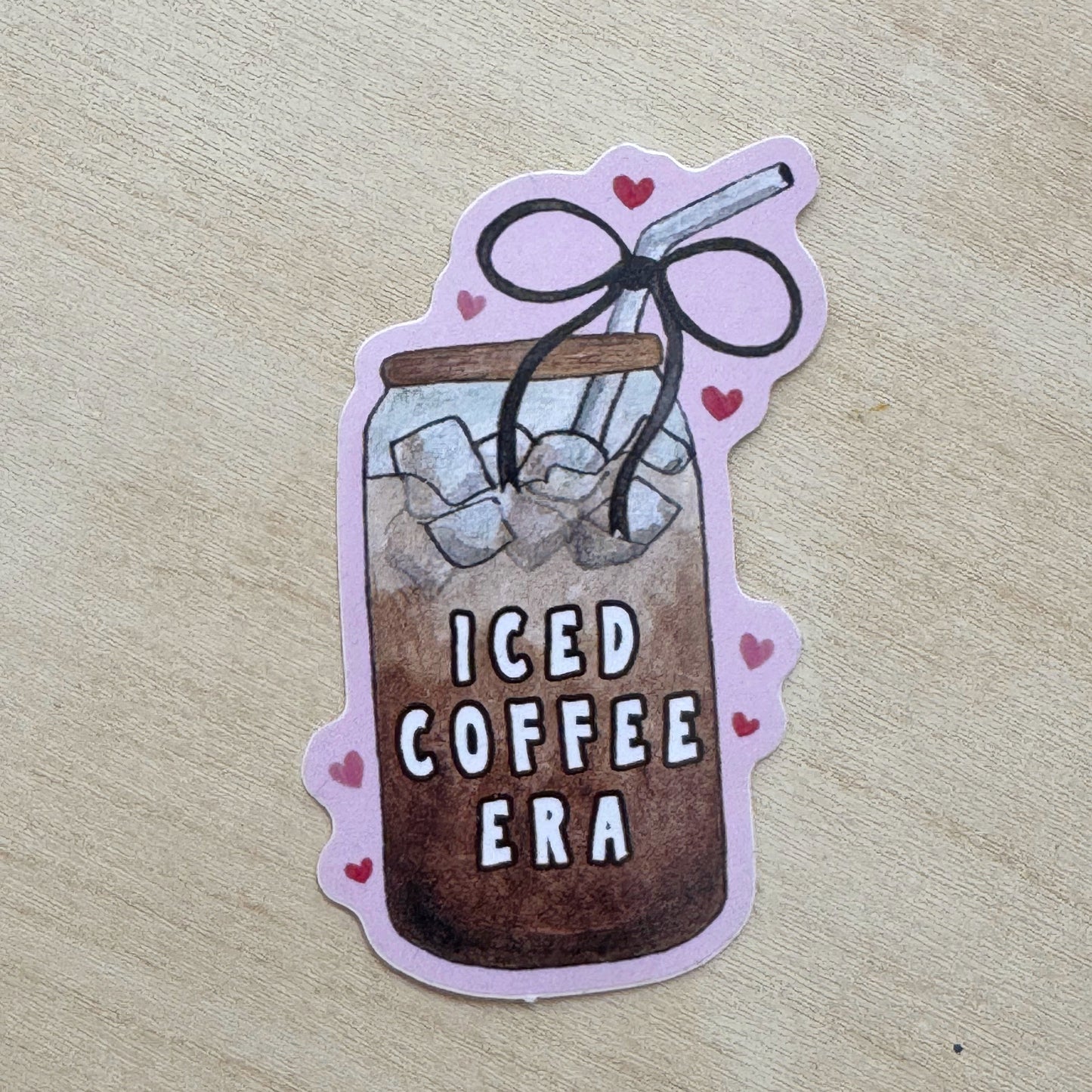A die-cut sticker featuring an illustration of an iced coffee in a glass jar with the text 'ICED COFFEE ERA' and small pink hearts, placed on a light wood surface.