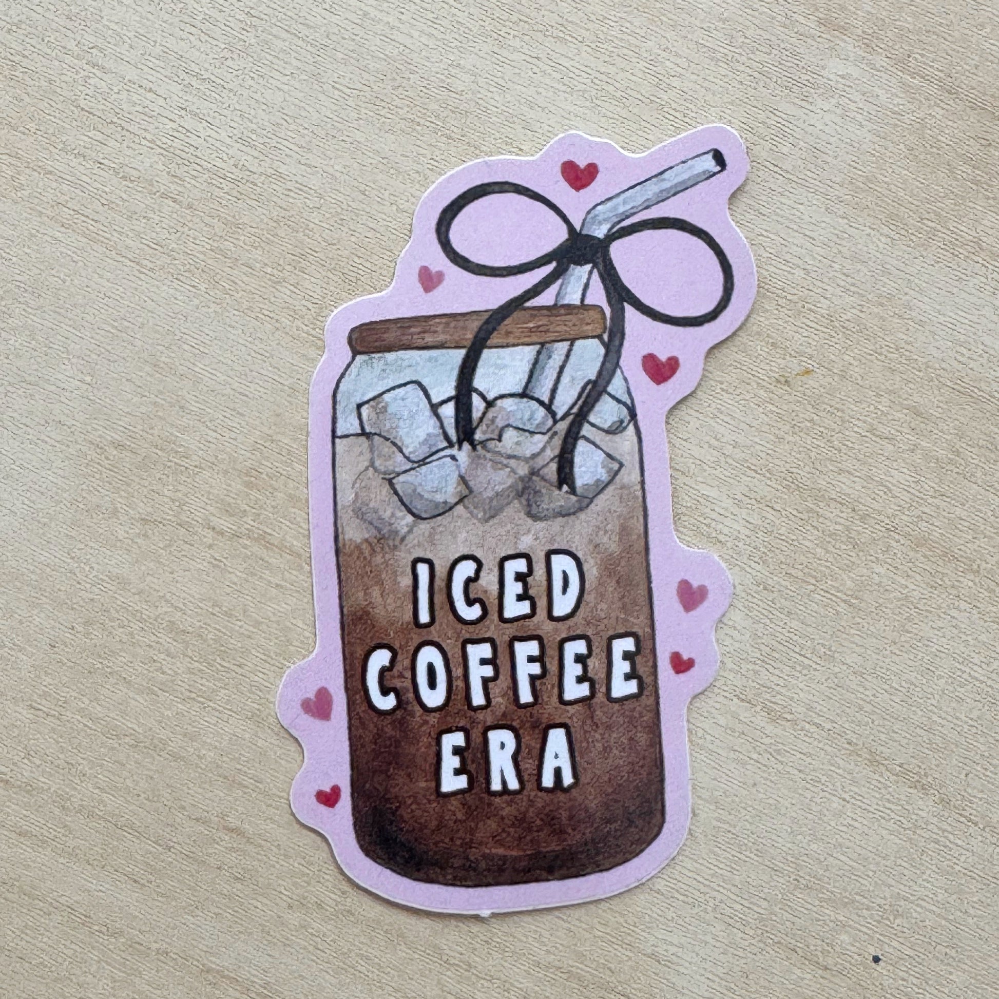 A die-cut sticker featuring an illustration of an iced coffee in a glass jar with the text 'ICED COFFEE ERA' and small pink hearts, placed on a light wood surface.